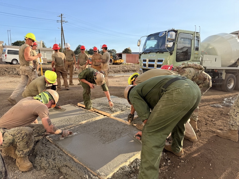A group of military personnel in uniform are actively pouring and finishing concrete slabs at a construction site. Workers wearing hard hats in yellow, red, and white are collaborating on the concrete work, with an Isuzu concrete mixer truck visible in the background. The scene appears to be a military engineering or construction exercise, likely a joint operation given the mix of uniform styles.