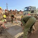 A group of military personnel in uniform are actively pouring and finishing concrete slabs at a construction site. Workers wearing hard hats in yellow, red, and white are collaborating on the concrete work, with an Isuzu concrete mixer truck visible in the background. The scene appears to be a military engineering or construction exercise, likely a joint operation given the mix of uniform styles.