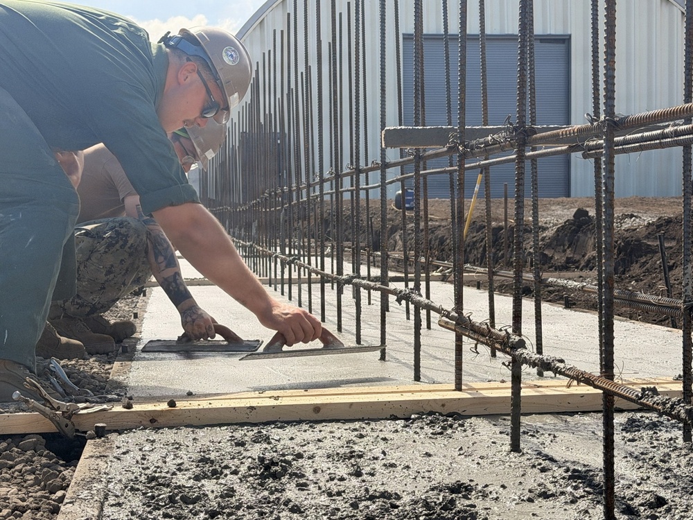 A construction worker wearing a hard hat, safety glasses, and military-style camouflage pants is finishing freshly poured concrete using a hand trowel. The work is being performed along a foundation or footing form bordered by numerous vertical rebar (reinforcing steel bars) that extend upward. A metal building structure is visible in the background of what appears to be an active construction site.