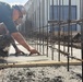 A construction worker wearing a hard hat, safety glasses, and military-style camouflage pants is finishing freshly poured concrete using a hand trowel. The work is being performed along a foundation or footing form bordered by numerous vertical rebar (reinforcing steel bars) that extend upward. A metal building structure is visible in the background of what appears to be an active construction site.