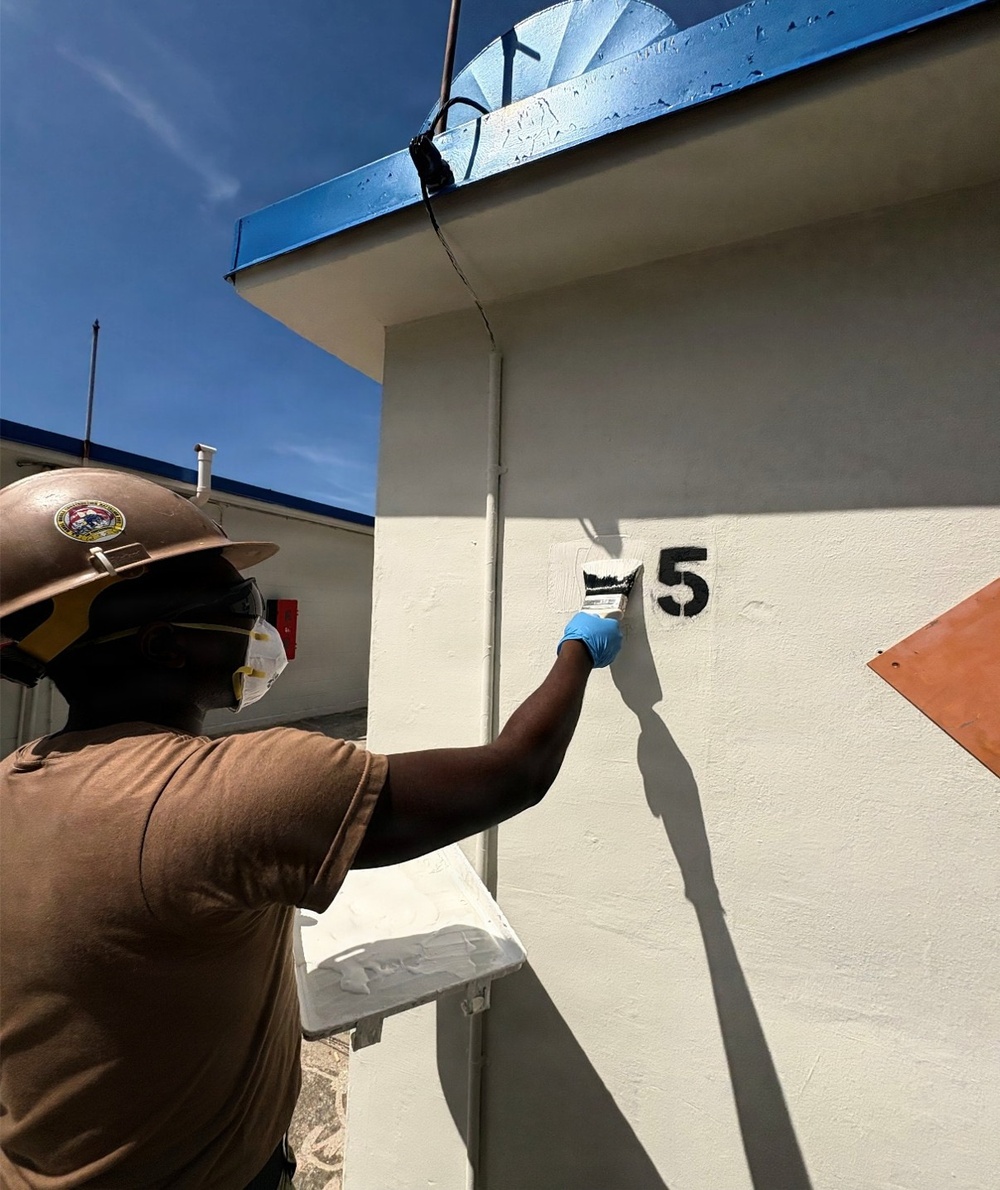 A worker wearing personal protective equipment — including a hard hat, face mask, and blue gloves — is painting a white exterior wall of a building. The worker is using a paintbrush and a small paint tray to apply white paint near a large black number '5' on the wall. The setting appears to be an outdoor industrial or institutional facility under clear blue skies.