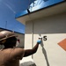 A worker wearing personal protective equipment — including a hard hat, face mask, and blue gloves — is painting a white exterior wall of a building. The worker is using a paintbrush and a small paint tray to apply white paint near a large black number '5' on the wall. The setting appears to be an outdoor industrial or institutional facility under clear blue skies.