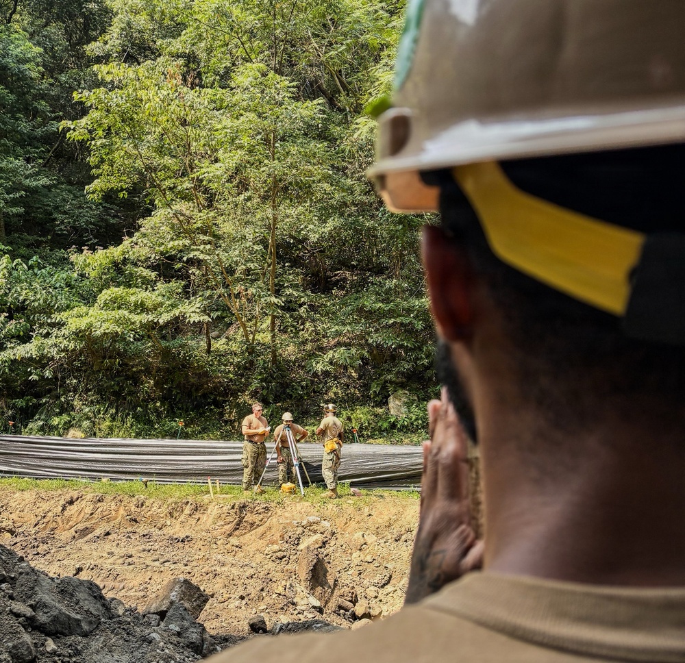 A construction or engineering site in a forested area where workers in military-style uniforms and hard hats are conducting surveying operations with a tripod-mounted instrument. A foreground observer wearing a tan hard hat with yellow markings watches the activity from an elevated position overlooking an excavated area. The setting appears to be a tropical or subtropical environment based on the dense green vegetation in the background.