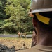 A construction or engineering site in a forested area where workers in military-style uniforms and hard hats are conducting surveying operations with a tripod-mounted instrument. A foreground observer wearing a tan hard hat with yellow markings watches the activity from an elevated position overlooking an excavated area. The setting appears to be a tropical or subtropical environment based on the dense green vegetation in the background.