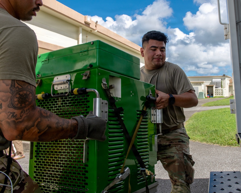 Two military personnel in OCP (Operational Camouflage Pattern) uniforms are handling a large green military equipment unit, likely a generator or power supply, on what appears to be a military installation. They are working together to move or position the heavy green metal equipment near a vehicle or loading platform. The setting appears to be an outdoor area of a military base with buildings visible in the background.