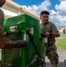 Two military personnel in OCP (Operational Camouflage Pattern) uniforms are handling a large green military equipment unit, likely a generator or power supply, on what appears to be a military installation. They are working together to move or position the heavy green metal equipment near a vehicle or loading platform. The setting appears to be an outdoor area of a military base with buildings visible in the background.