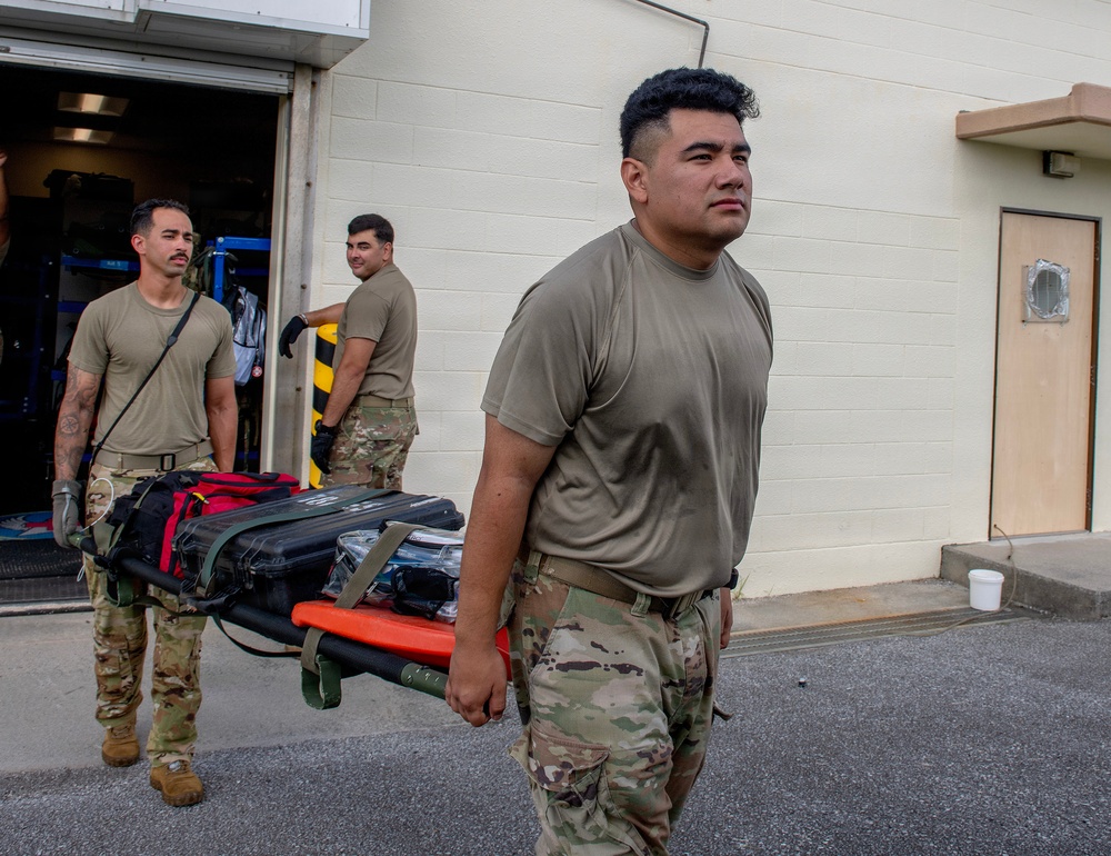 Three military personnel in OCP (Operational Camouflage Pattern) uniforms are handling medical equipment outside a building. Two individuals are carrying a stretcher loaded with medical bags and cases, while a third is unloading or loading equipment from what appears to be an ambulance or medical vehicle. The scene suggests a medical readiness exercise, deployment preparation, or emergency response operation.