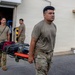 Three military personnel in OCP (Operational Camouflage Pattern) uniforms are handling medical equipment outside a building. Two individuals are carrying a stretcher loaded with medical bags and cases, while a third is unloading or loading equipment from what appears to be an ambulance or medical vehicle. The scene suggests a medical readiness exercise, deployment preparation, or emergency response operation.
