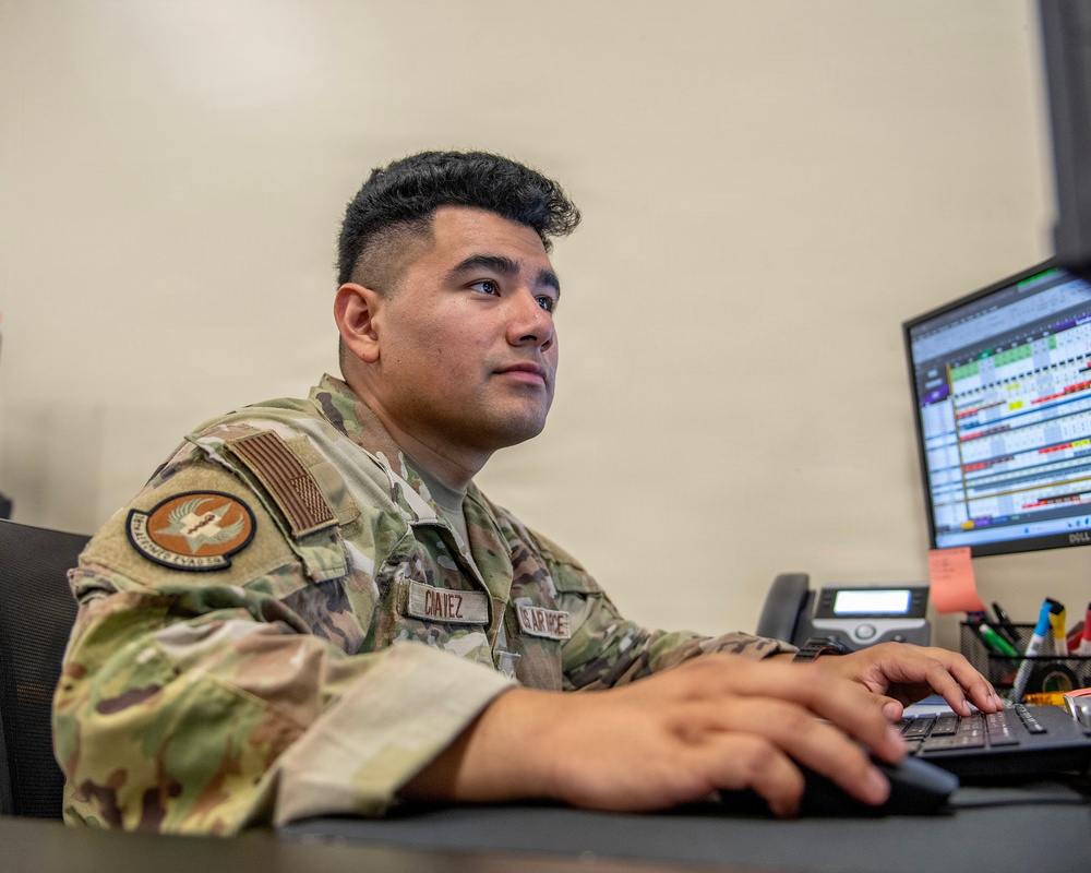 A U.S. Air Force service member identified by the name tape 'CHAVEZ' is seated at a desk working at a computer workstation. The individual is wearing an Operational Camouflage Pattern (OCP) uniform with a U.S. flag patch and a unit patch on the left sleeve. The monitor displays what appears to be a scheduling or data management spreadsheet.