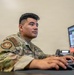 A U.S. Air Force service member identified by the name tape 'CHAVEZ' is seated at a desk working at a computer workstation. The individual is wearing an Operational Camouflage Pattern (OCP) uniform with a U.S. flag patch and a unit patch on the left sleeve. The monitor displays what appears to be a scheduling or data management spreadsheet.