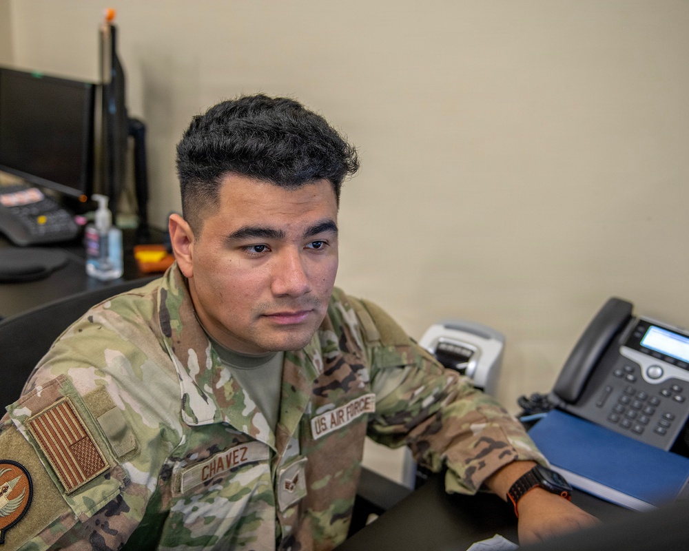 A U.S. Air Force servicemember named Chavez is seated at a desk in what appears to be an office or administrative workspace. He is wearing an Operational Camouflage Pattern (OCP) uniform with a U.S. flag patch on the left sleeve and a name tape reading 'CHAVEZ' on the chest. The image is a close-up portrait-style photograph taken in an indoor military office environment.