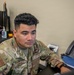 A U.S. Air Force servicemember named Chavez is seated at a desk in what appears to be an office or administrative workspace. He is wearing an Operational Camouflage Pattern (OCP) uniform with a U.S. flag patch on the left sleeve and a name tape reading 'CHAVEZ' on the chest. The image is a close-up portrait-style photograph taken in an indoor military office environment.