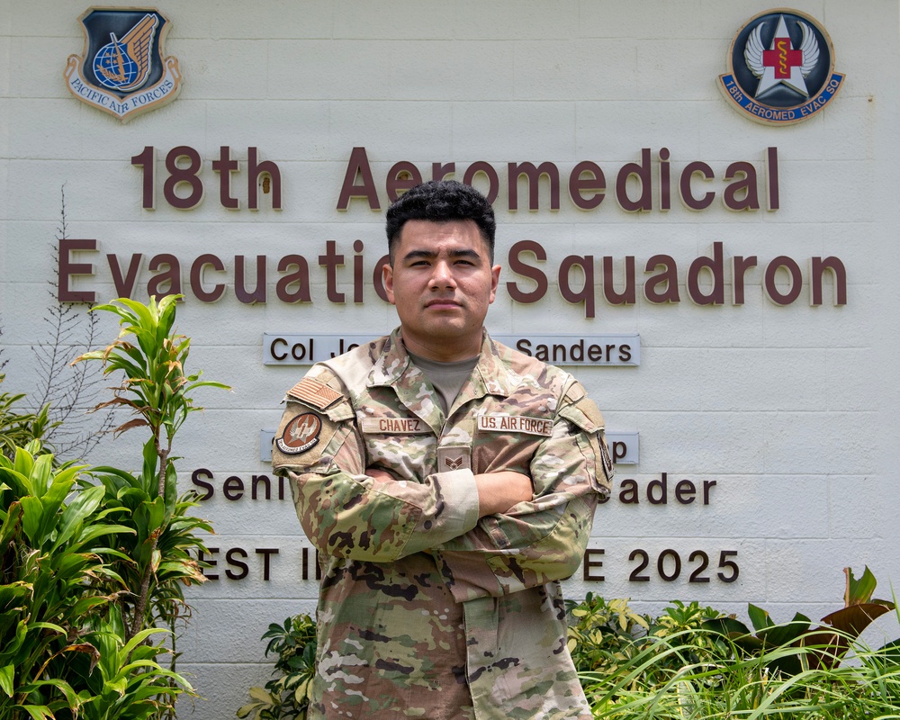A U.S. Air Force service member named Chavez stands with arms crossed in front of the 18th Aeromedical Evacuation Squadron building sign. The individual is wearing an OCP (Operational Camouflage Pattern) uniform with visible name tape, U.S. Air Force tape, and rank insignia. The setting appears to be at a military installation, likely in the Pacific region based on the Pacific Air Forces emblem visible on the sign.