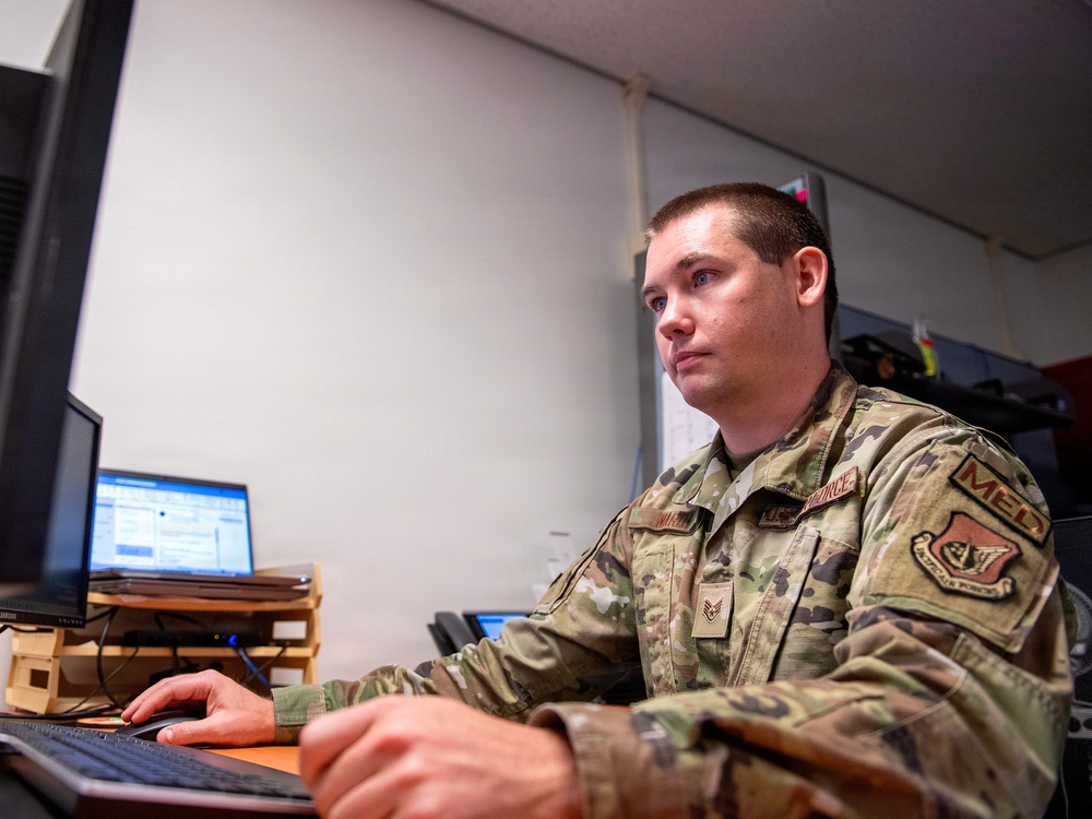 A U.S. Air Force enlisted member in Operational Camouflage Pattern (OCP) uniform is seated at a desk working at a computer workstation. The individual displays Staff Sergeant rank insignia and wears a 'MED' patch on the right shoulder, indicating a medical unit assignment. The setting appears to be a military office or administrative workspace.