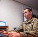 A U.S. Air Force enlisted member in Operational Camouflage Pattern (OCP) uniform is seated at a desk working at a computer workstation. The individual displays Staff Sergeant rank insignia and wears a 'MED' patch on the right shoulder, indicating a medical unit assignment. The setting appears to be a military office or administrative workspace.