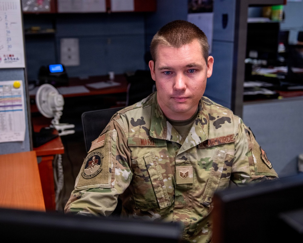 A U.S. Air Force Staff Sergeant named Ward is seated at a desk in an office environment, wearing Operational Camouflage Pattern (OCP) uniform. He is looking directly at the camera with a neutral expression. The setting appears to be a military administrative or medical support office.