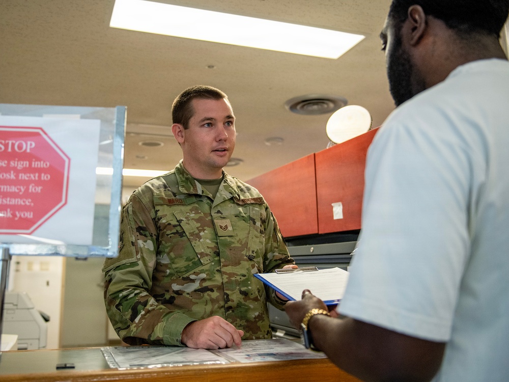 A U.S. Air Force service member in OCP (Operational Camouflage Pattern) uniform is speaking with a civilian at what appears to be a military pharmacy or administrative counter. The airman is holding a blue clipboard with paperwork while engaging with the civilian patron. A red octagonal 'STOP' sign with pharmacy check-in instructions is visible in the background.
