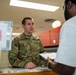 A U.S. Air Force service member in OCP (Operational Camouflage Pattern) uniform is speaking with a civilian at what appears to be a military pharmacy or administrative counter. The airman is holding a blue clipboard with paperwork while engaging with the civilian patron. A red octagonal 'STOP' sign with pharmacy check-in instructions is visible in the background.