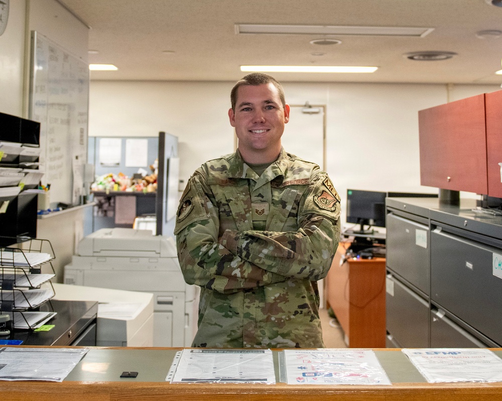 A U.S. Air Force service member in Operational Camouflage Pattern (OCP) uniform stands with arms crossed behind a service counter, smiling at the camera. The setting appears to be a military administrative office, likely a personnel or support services office based on visible signage including 'EFMP' (Exceptional Family Member Program) brochures on the counter. The environment includes office equipment, filing cabinets, and a whiteboard in the background.