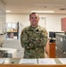 A U.S. Air Force service member in Operational Camouflage Pattern (OCP) uniform stands with arms crossed behind a service counter, smiling at the camera. The setting appears to be a military administrative office, likely a personnel or support services office based on visible signage including 'EFMP' (Exceptional Family Member Program) brochures on the counter. The environment includes office equipment, filing cabinets, and a whiteboard in the background.