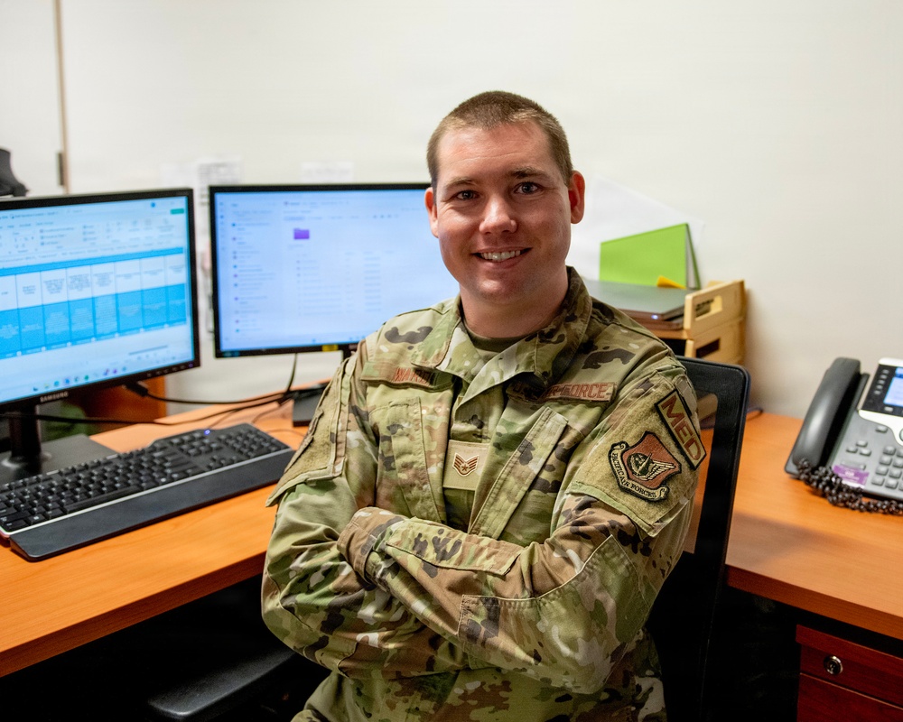 A U.S. Air Force enlisted member in OCP (Operational Camouflage Pattern) uniform sits at a dual-monitor workstation with arms crossed, smiling at the camera. The individual displays rank insignia consistent with a Staff Sergeant and wears patches including 'U.S. AIR FORCE' tape and what appears to be an 'MBD' unit patch. The setting appears to be a military administrative or operations office environment.