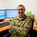 A U.S. Air Force enlisted member in OCP (Operational Camouflage Pattern) uniform sits at a dual-monitor workstation with arms crossed, smiling at the camera. The individual displays rank insignia consistent with a Staff Sergeant and wears patches including 'U.S. AIR FORCE' tape and what appears to be an 'MBD' unit patch. The setting appears to be a military administrative or operations office environment.