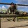A female U.S. Marine in digital camouflage (MARPAT) uniform is navigating a wooden log obstacle course, performing a split-like maneuver over parallel horizontal logs. The setting appears to be a military training facility with a sandy ground surface and open sky with scattered clouds. The image captures an active physical training exercise on what appears to be a standard military obstacle course.