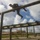 A soldier in military camouflage uniform and combat boots is navigating a wooden obstacle course, captured mid-motion as they vault over a horizontal log beam. The image is taken from a low angle, emphasizing the height of the obstacle against a partly cloudy sky. Military installation buildings are visible in the background, confirming this is a training facility.