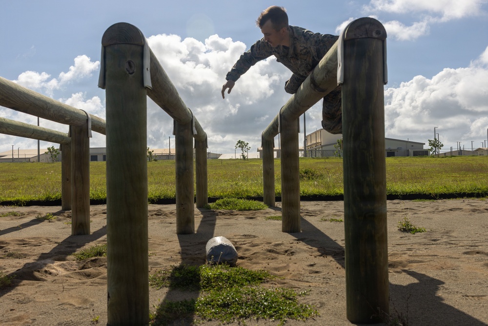 A person in military camouflage uniform is navigating a wooden obstacle course, appearing to vault or climb over a horizontal log barrier. The obstacle course is set on a sandy surface with military-style buildings visible in the background. The image is taken from a low angle, emphasizing the wooden posts and horizontal rails of the obstacle structure.