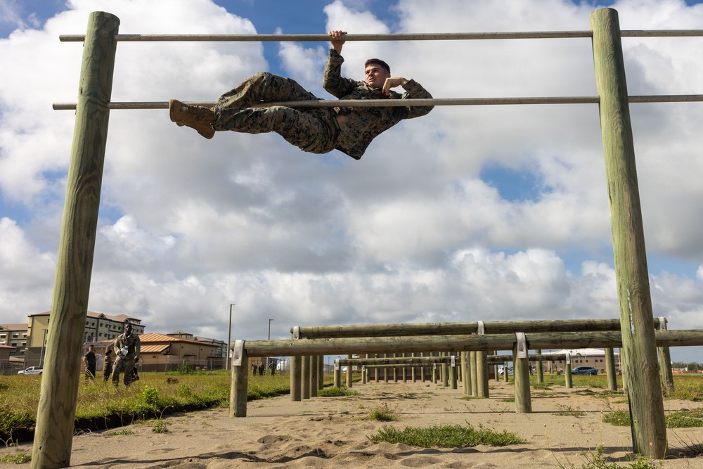 A military service member in camouflage uniform is navigating a horizontal bar obstacle course, performing an advanced body maneuver with legs raised and body parallel to the bar. The setting appears to be a military base obstacle/fitness training course with multiple wooden log and metal bar structures extending into the background. Other personnel in uniform are visible in the background observing or waiting their turn.