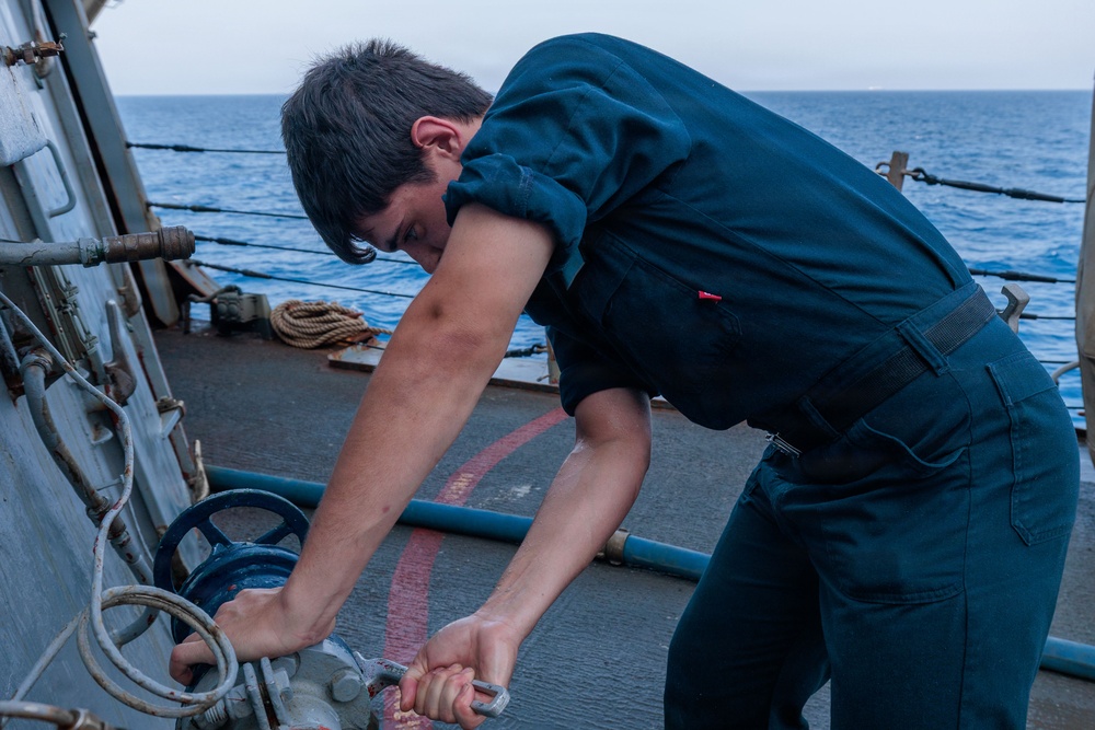 A young male sailor in dark navy coveralls is working on deck equipment aboard a naval vessel at sea. He is bent over, applying force to what appears to be a valve or fitting using a wrench or tool. The ocean is visible in the background under overcast skies.