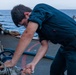 A young male sailor in dark navy coveralls is working on deck equipment aboard a naval vessel at sea. He is bent over, applying force to what appears to be a valve or fitting using a wrench or tool. The ocean is visible in the background under overcast skies.