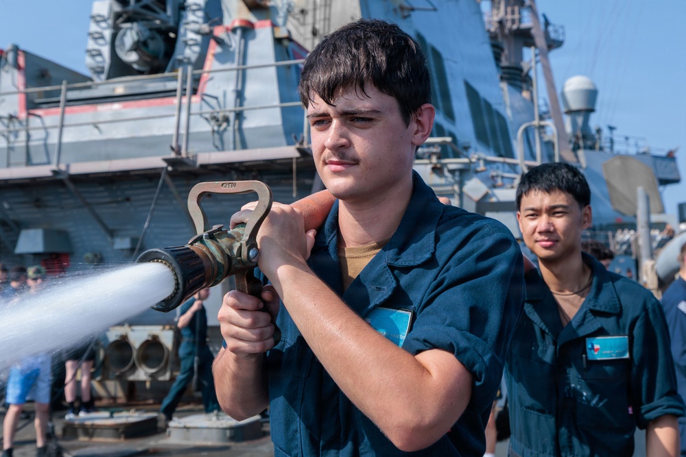 A young sailor in dark blue coveralls operates a fire hose nozzle on the deck of a naval vessel, with water actively spraying from the nozzle. A second sailor in matching coveralls stands behind him observing. The setting appears to be a damage control or firefighting drill conducted aboard a warship.