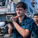A young sailor in dark blue coveralls operates a fire hose nozzle on the deck of a naval vessel, with water actively spraying from the nozzle. A second sailor in matching coveralls stands behind him observing. The setting appears to be a damage control or firefighting drill conducted aboard a warship.