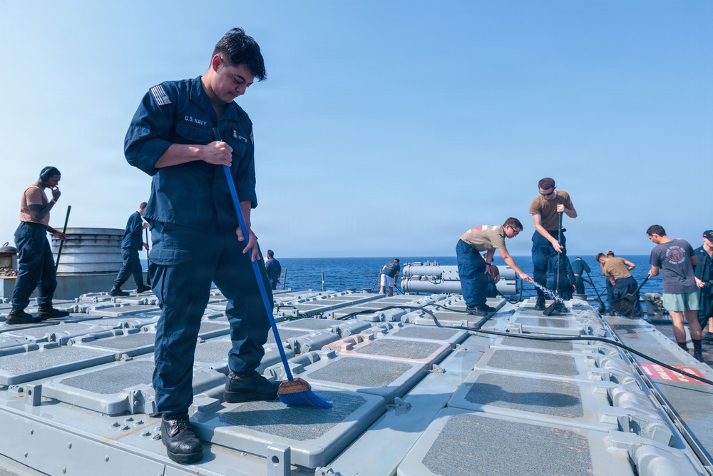 U.S. Navy sailors are performing routine deck maintenance and cleaning operations aboard a naval vessel at sea. The primary subject in the foreground is wearing a blue U.S. Navy uniform and sweeping the deck with a blue-handled broom. Multiple crew members in various uniforms are visible across the deck, engaged in similar maintenance tasks.