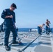 U.S. Navy sailors are performing routine deck maintenance and cleaning operations aboard a naval vessel at sea. The primary subject in the foreground is wearing a blue U.S. Navy uniform and sweeping the deck with a blue-handled broom. Multiple crew members in various uniforms are visible across the deck, engaged in similar maintenance tasks.