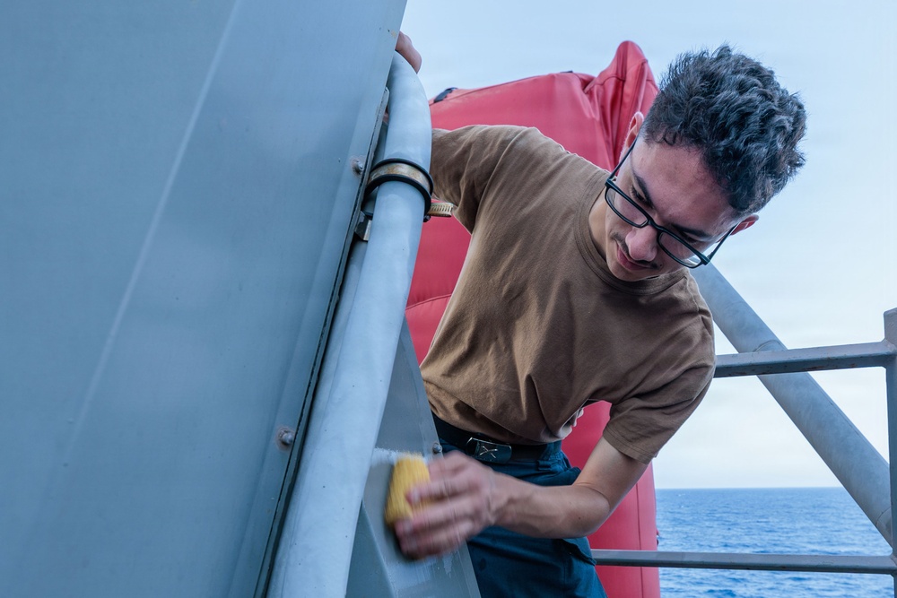 A young man wearing glasses and a brown t-shirt is scrubbing or cleaning a metal railing or surface on what appears to be a naval vessel or ship. He is using a yellow sponge or cleaning pad and is leaning over the railing to reach the surface. The ocean is visible in the background, confirming the maritime setting.