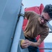 A young man wearing glasses and a brown t-shirt is scrubbing or cleaning a metal railing or surface on what appears to be a naval vessel or ship. He is using a yellow sponge or cleaning pad and is leaning over the railing to reach the surface. The ocean is visible in the background, confirming the maritime setting.