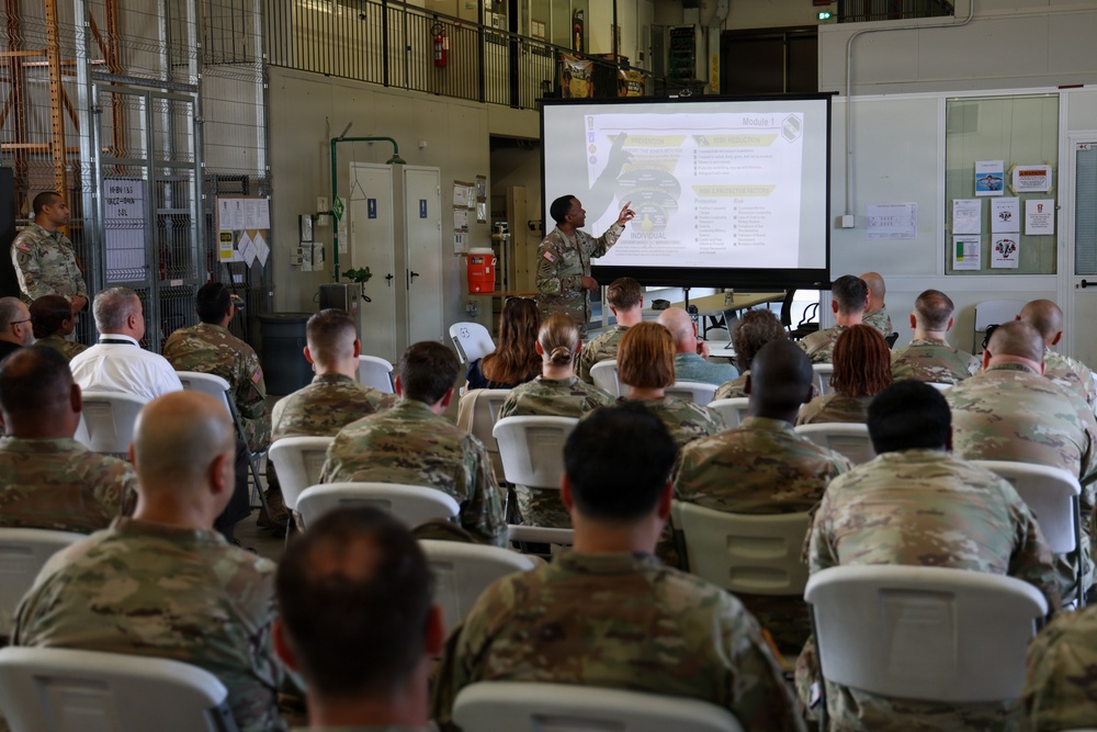A military briefing or training session is taking place in what appears to be a warehouse or motor pool facility. A soldier is presenting at the front of the room, pointing to a projected slide labeled 'Module 1' to an audience of approximately 30-40 military personnel seated in white folding chairs. The audience consists primarily of soldiers in OCP (Operational Camouflage Pattern) uniforms, with a few civilians also present.