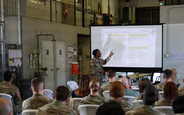 A military briefing or training session is taking place in what appears to be a warehouse or motor pool facility. A soldier is presenting at the front of the room, pointing to a projected slide labeled 'Module 1' to an audience of approximately 30-40 military personnel seated in white folding chairs. The audience consists primarily of soldiers in OCP (Operational Camouflage Pattern) uniforms, with a few civilians also present.