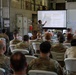A military briefing or training session is taking place in what appears to be a warehouse or motor pool facility. A soldier is presenting at the front of the room, pointing to a projected slide labeled 'Module 1' to an audience of approximately 30-40 military personnel seated in white folding chairs. The audience consists primarily of soldiers in OCP (Operational Camouflage Pattern) uniforms, with a few civilians also present.