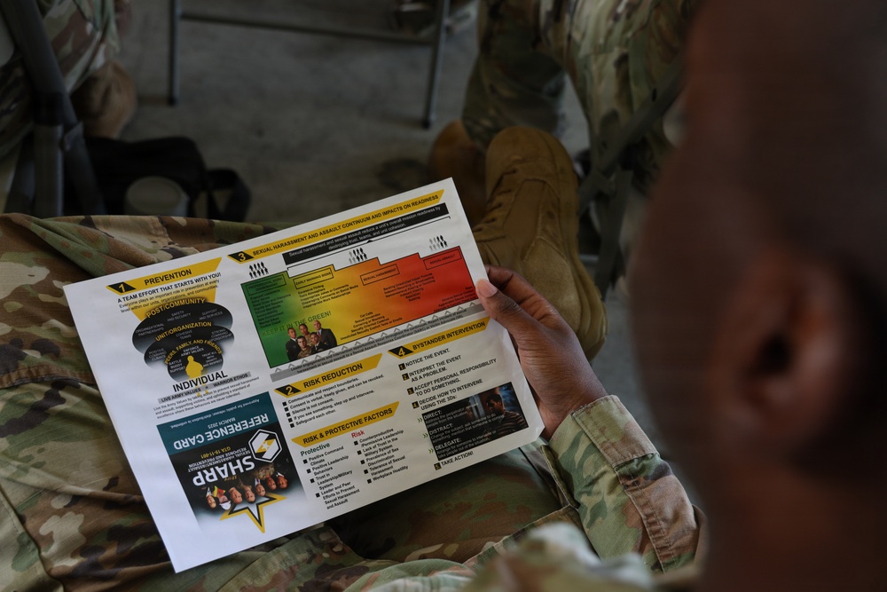 A soldier in camouflage uniform is reading a SHARP (Sexual Harassment/Assault Response and Prevention) Reference Card. The document appears to be an official U.S. Army training material covering prevention, risk reduction, bystander intervention, and related topics. The image is taken from an over-the-shoulder perspective during what appears to be a training or briefing session.