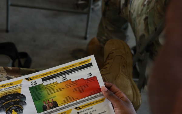 A soldier in camouflage uniform is reading a SHARP (Sexual Harassment/Assault Response and Prevention) Reference Card. The document appears to be an official U.S. Army training material covering prevention, risk reduction, bystander intervention, and related topics. The image is taken from an over-the-shoulder perspective during what appears to be a training or briefing session.