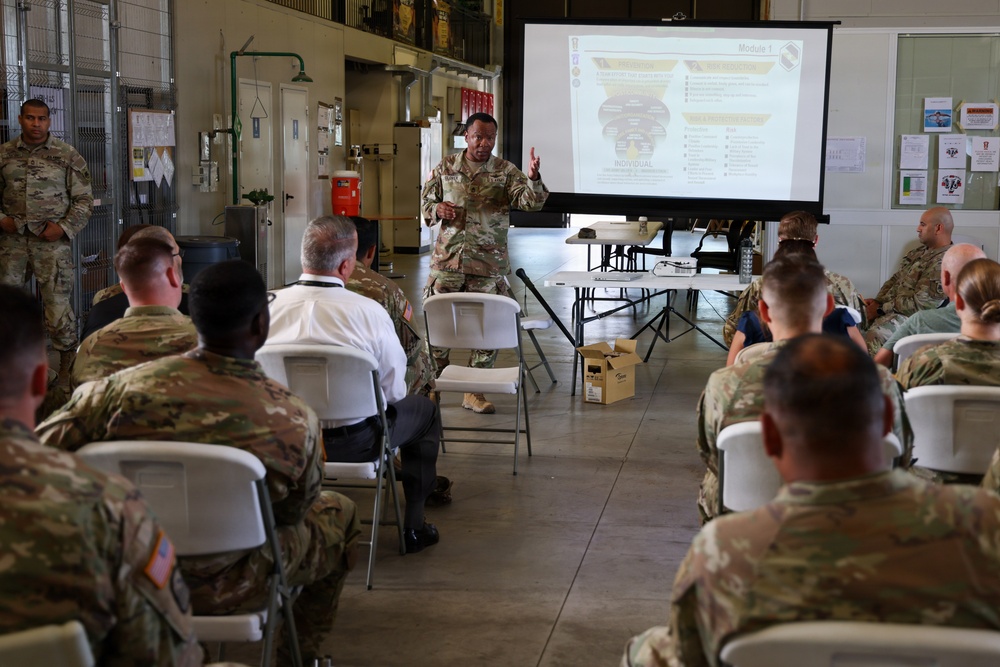 A military briefing or training session is taking place in what appears to be a hangar or maintenance bay facility. A soldier in camouflage uniform is presenting at the front of the room using a projected slideshow labeled 'Module 1,' while approximately 15-20 military personnel and at least one civilian are seated in white folding chairs. The setting suggests a formal instructional or leadership development event.
