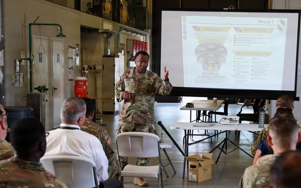 A military briefing or training session is taking place in what appears to be a hangar or maintenance bay facility. A soldier in camouflage uniform is presenting at the front of the room using a projected slideshow labeled 'Module 1,' while approximately 15-20 military personnel and at least one civilian are seated in white folding chairs. The setting suggests a formal instructional or leadership development event.