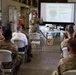 A military briefing or training session is taking place in what appears to be a hangar or maintenance bay facility. A soldier in camouflage uniform is presenting at the front of the room using a projected slideshow labeled 'Module 1,' while approximately 15-20 military personnel and at least one civilian are seated in white folding chairs. The setting suggests a formal instructional or leadership development event.