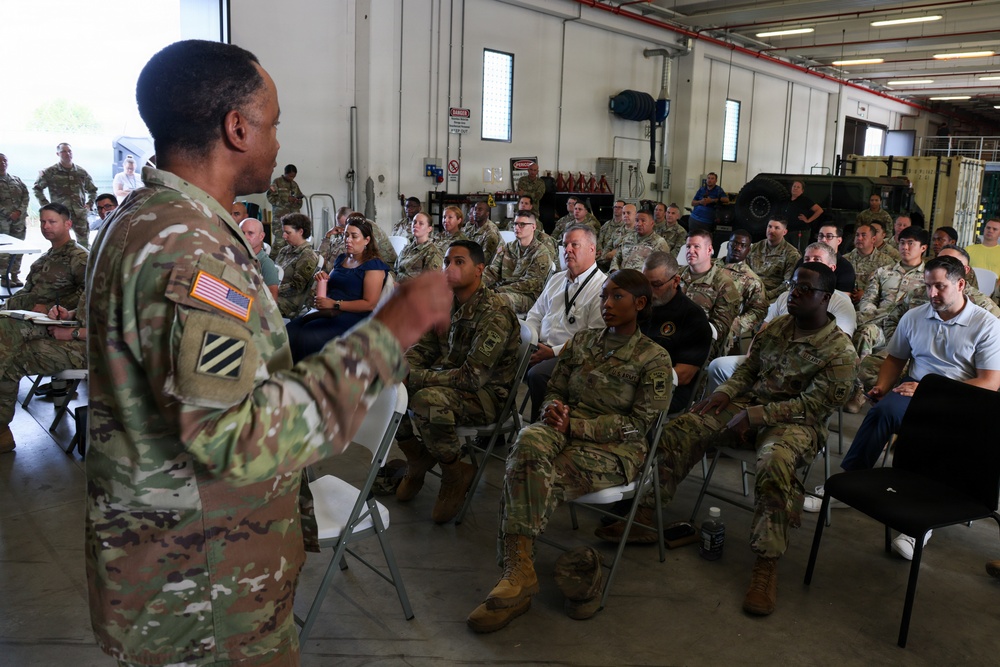 A U.S. Army soldier wearing OCP (Operational Camouflage Pattern) uniform with a 3rd Infantry Division patch and American flag is addressing a large group of military personnel and some civilians seated in folding chairs inside what appears to be a military maintenance bay or motor pool facility. The audience consists primarily of soldiers in OCP uniforms along with a few civilians in business casual attire. The setting suggests a formal briefing, town hall, or command information session.
