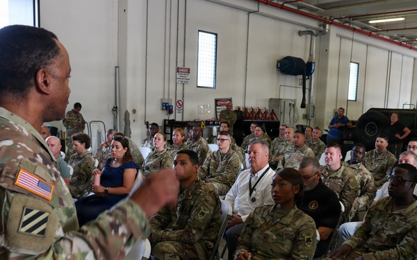 A U.S. Army soldier wearing OCP (Operational Camouflage Pattern) uniform with a 3rd Infantry Division patch and American flag is addressing a large group of military personnel and some civilians seated in folding chairs inside what appears to be a military maintenance bay or motor pool facility. The audience consists primarily of soldiers in OCP uniforms along with a few civilians in business casual attire. The setting suggests a formal briefing, town hall, or command information session.
