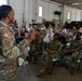A U.S. Army soldier wearing OCP (Operational Camouflage Pattern) uniform with a 3rd Infantry Division patch and American flag is addressing a large group of military personnel and some civilians seated in folding chairs inside what appears to be a military maintenance bay or motor pool facility. The audience consists primarily of soldiers in OCP uniforms along with a few civilians in business casual attire. The setting suggests a formal briefing, town hall, or command information session.
