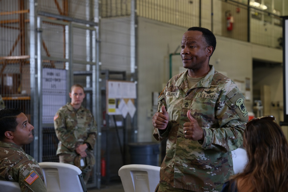 A U.S. Army soldier named Glover, identifiable by name tape and rank insignia, appears to be addressing a small group in what looks like a military hangar or warehouse facility. He is gesturing with both hands in a thumbs-up position, suggesting an instructional or motivational briefing. At least two other uniformed soldiers are visible — one seated and one standing in the background.